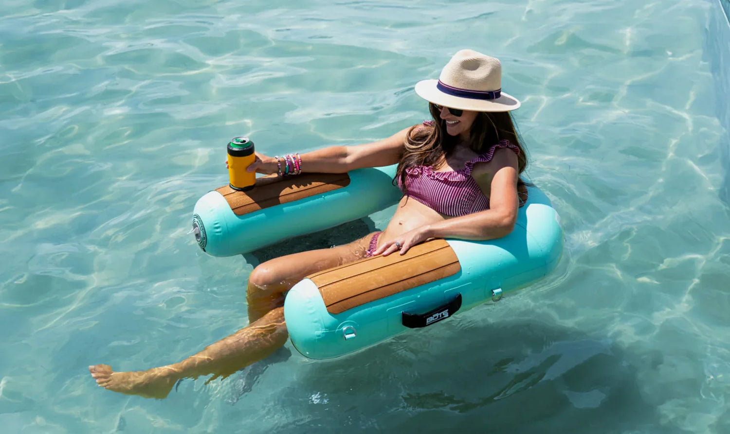 Person relaxing on a float in clear water, wearing a hat and sipping a drink.