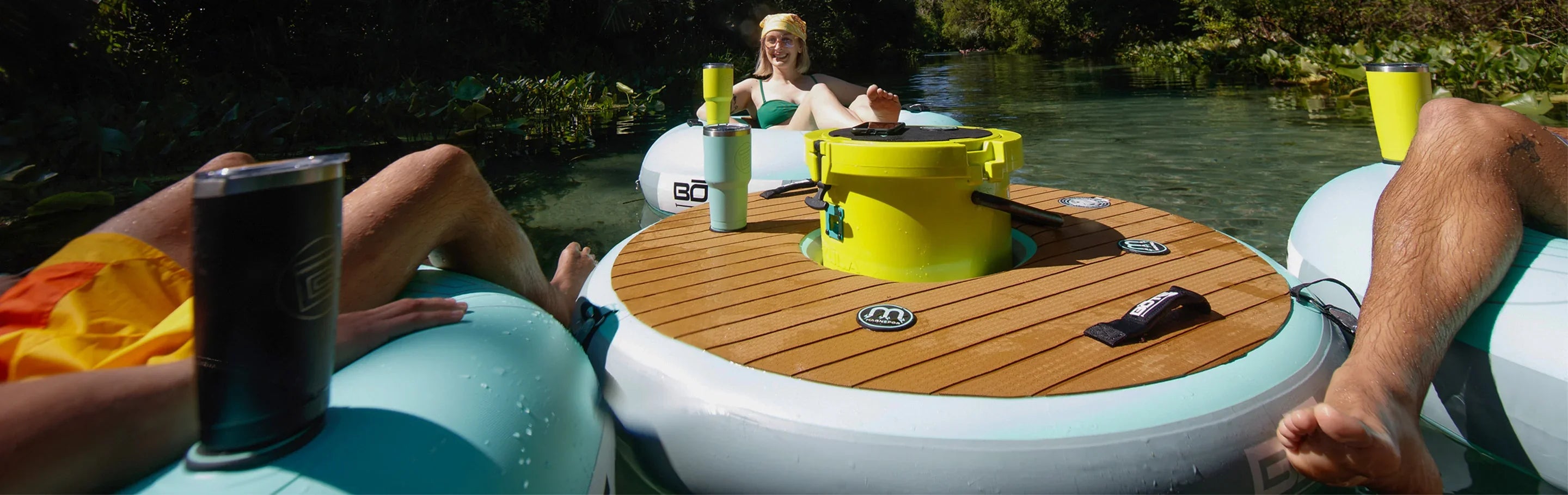 People relaxing on inflatable tube seats around the Bote Float, a floating table with a built-in cooler, cup holders, and a wood-like deck, floating on calm water.