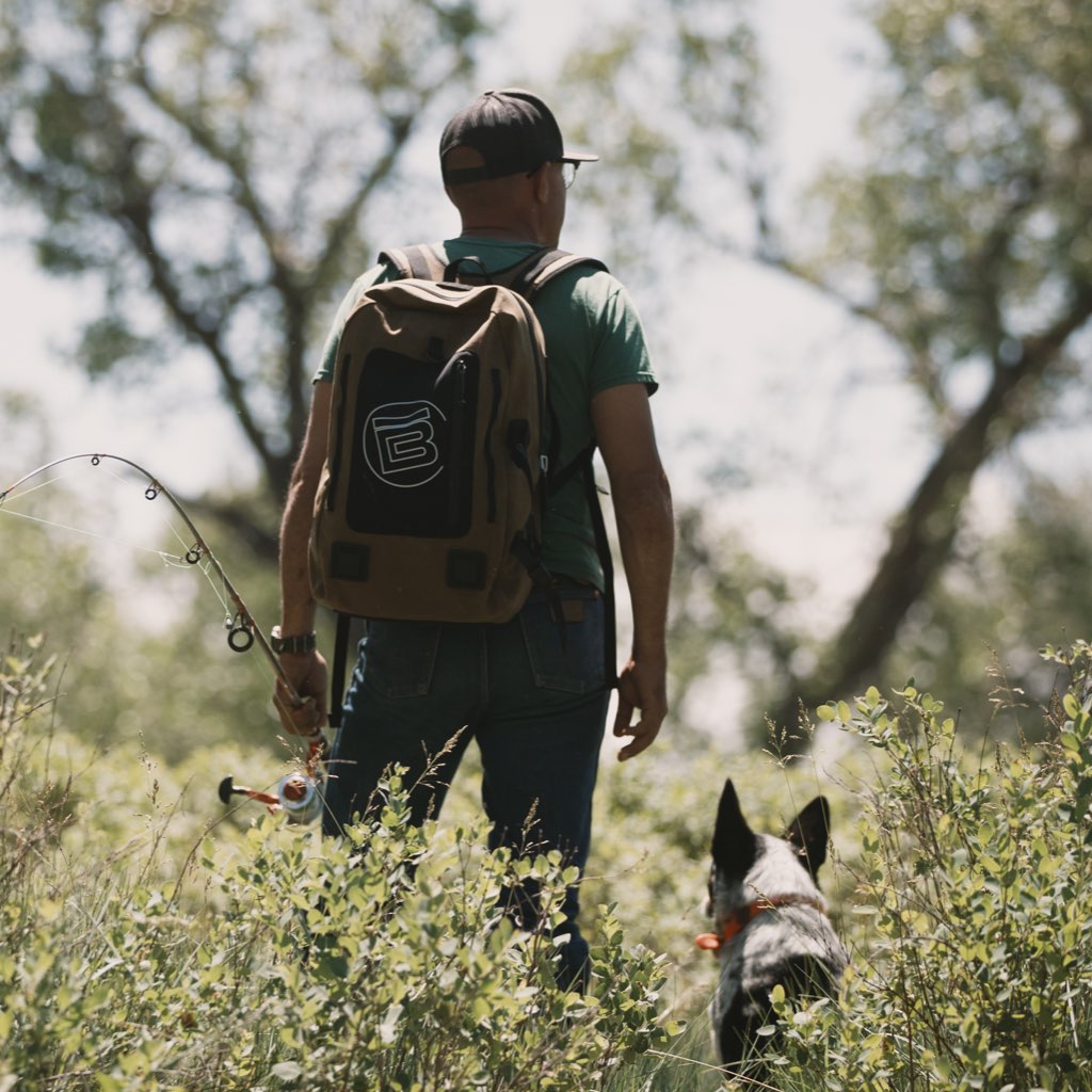 Man hiking with Highwater Backpack Last Cowboy while holding a fishing pole, dog beside him.