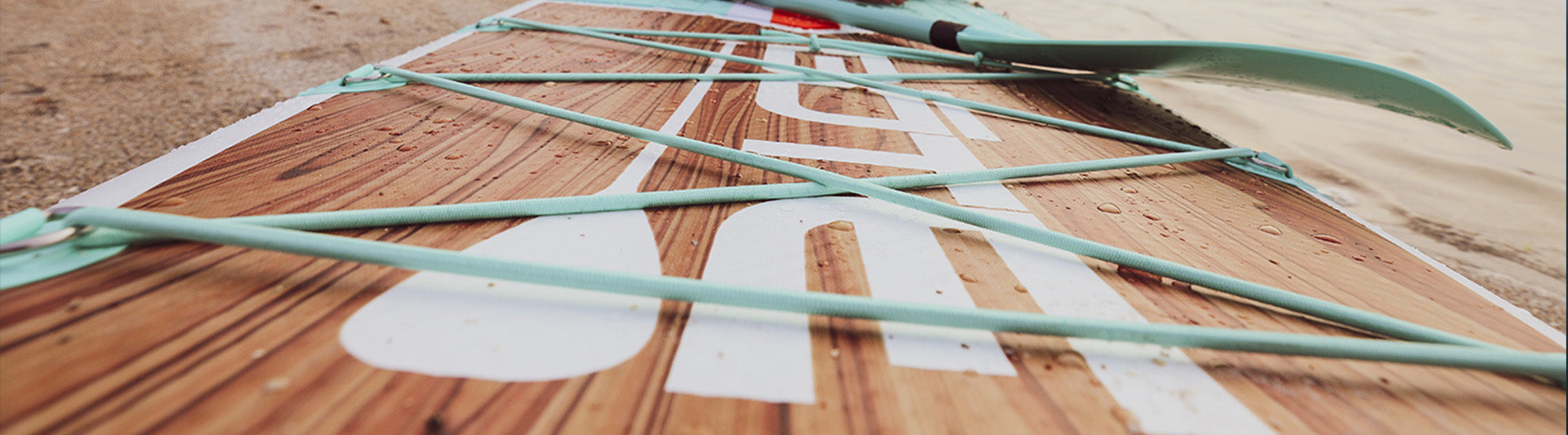 Close-up of woodgrain paddle board with mint bungee cords resting on the sand.