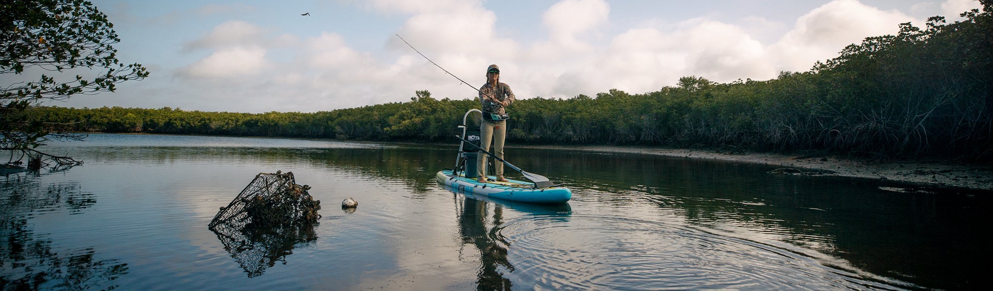 Woman fishing from blue inflatable paddle board near trees on calm water.