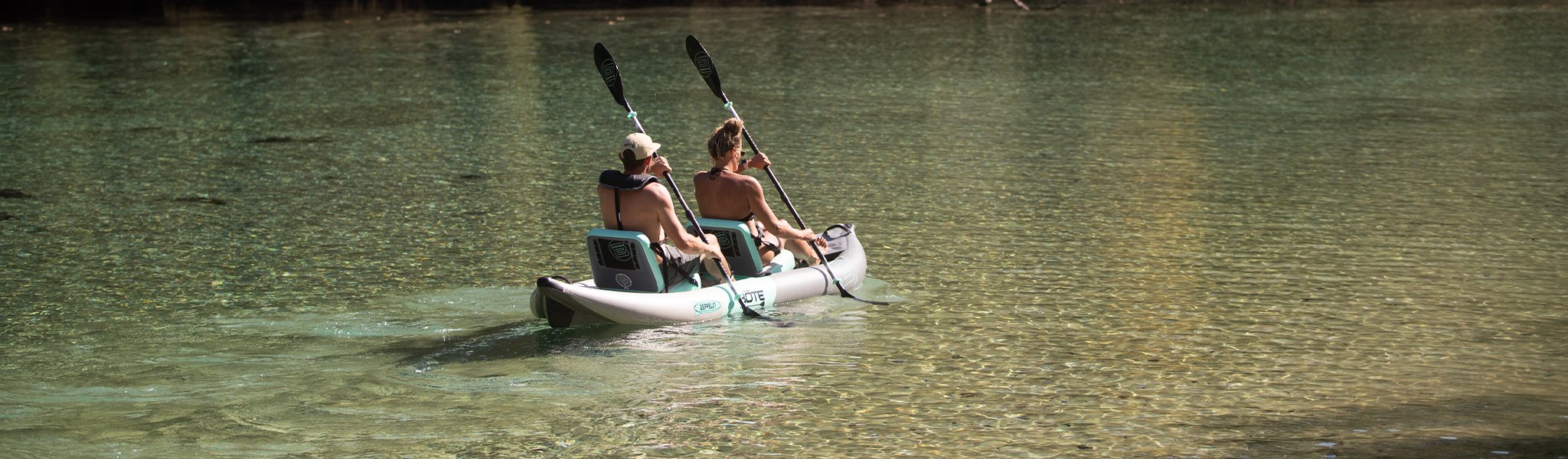 Two people paddling tandem inflatable kayak across clear shallow water.