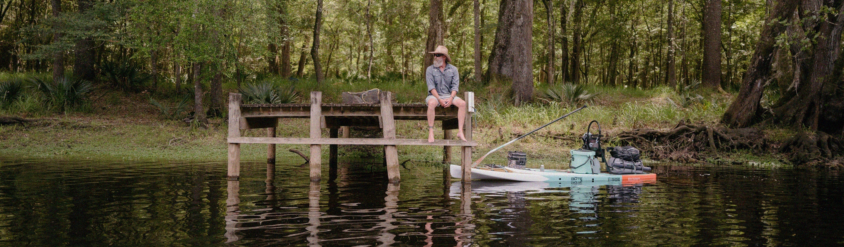 Person sitting on a wooden dock by a lake with a kayak nearby, surrounded by trees.