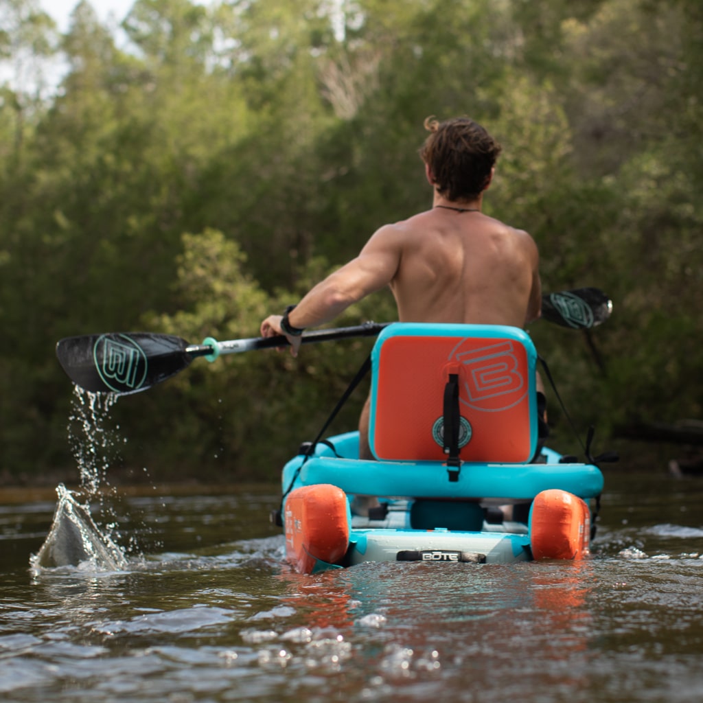 Person paddling a blue and orange kayak on water with trees in the background